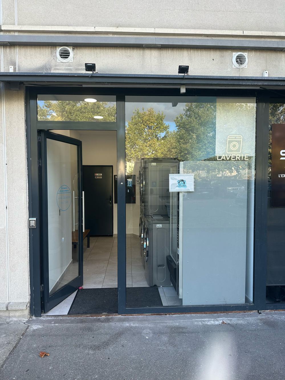 Devanture de la laverie : porte vitrée anthracite, enseigne lumineuse LAVERIE et machines visibles derrière la vitrine